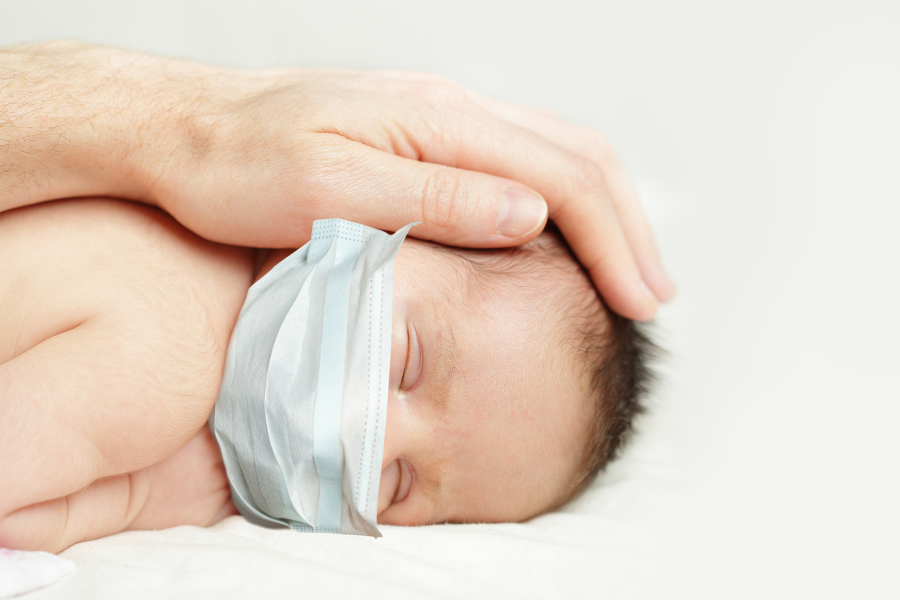 Newborn baby wearing a surgical mask with a parents hand gently holding it's head