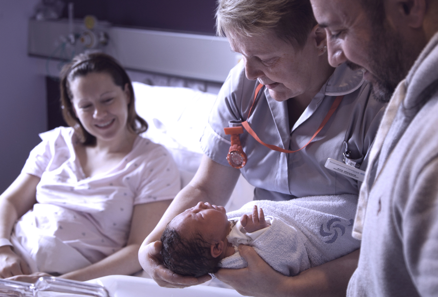 Midwife holds a newborn baby while new mum looks on from a hospital bed and dad stands closeby