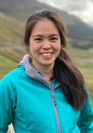 A smiling Asian woman standing in front of a hill which is blurred in the background. Her long dark brown hair is in a ponytail over her right shoulder and she is wearing a warm turquoise jacket.